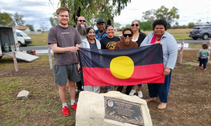 People gathered at Wales Park in Moree hold the Aboriginal flag above the memorial stone for Aunty Heather Boney, unveiled on the 25th anniversary of her death.