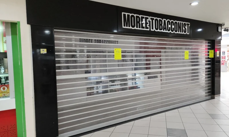 Closed roller shutter beneath the MOREE TOBACCONIST sign at Balo Square Shopping Centre, with yellow closure notices on the shutter and the right-hand black pillar.